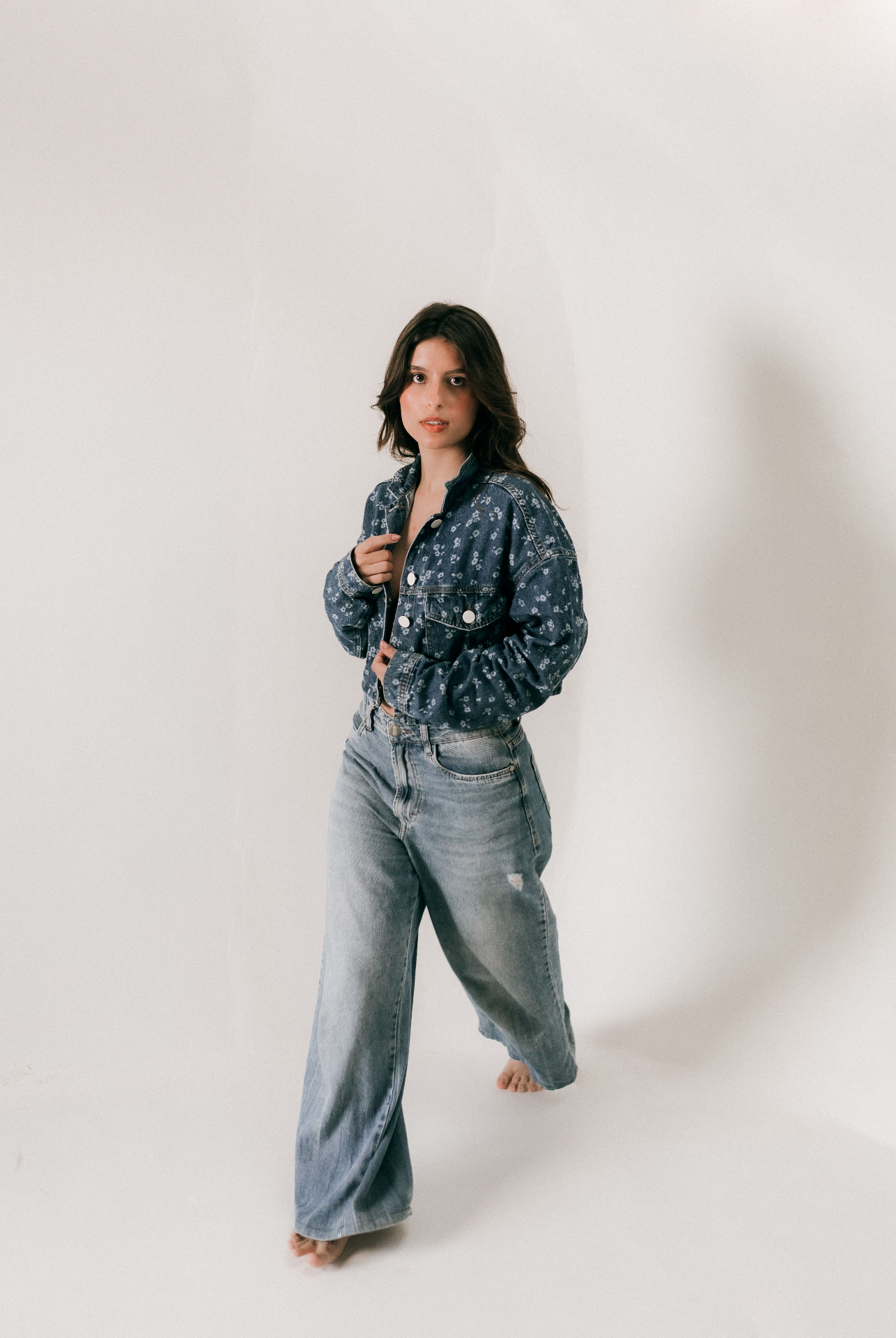 Woman in a cropped floral-print denim jacket with light-wash wide-leg jeans in a bright studio