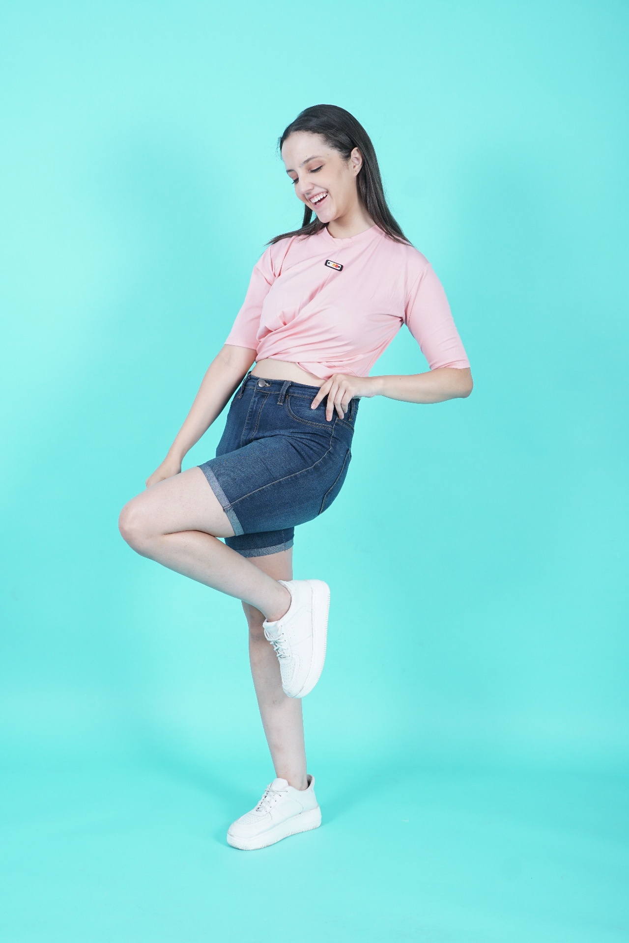 Woman in pink knotted crop tee and dark-wash mid-thigh denim shorts on a turquoise studio backdrop