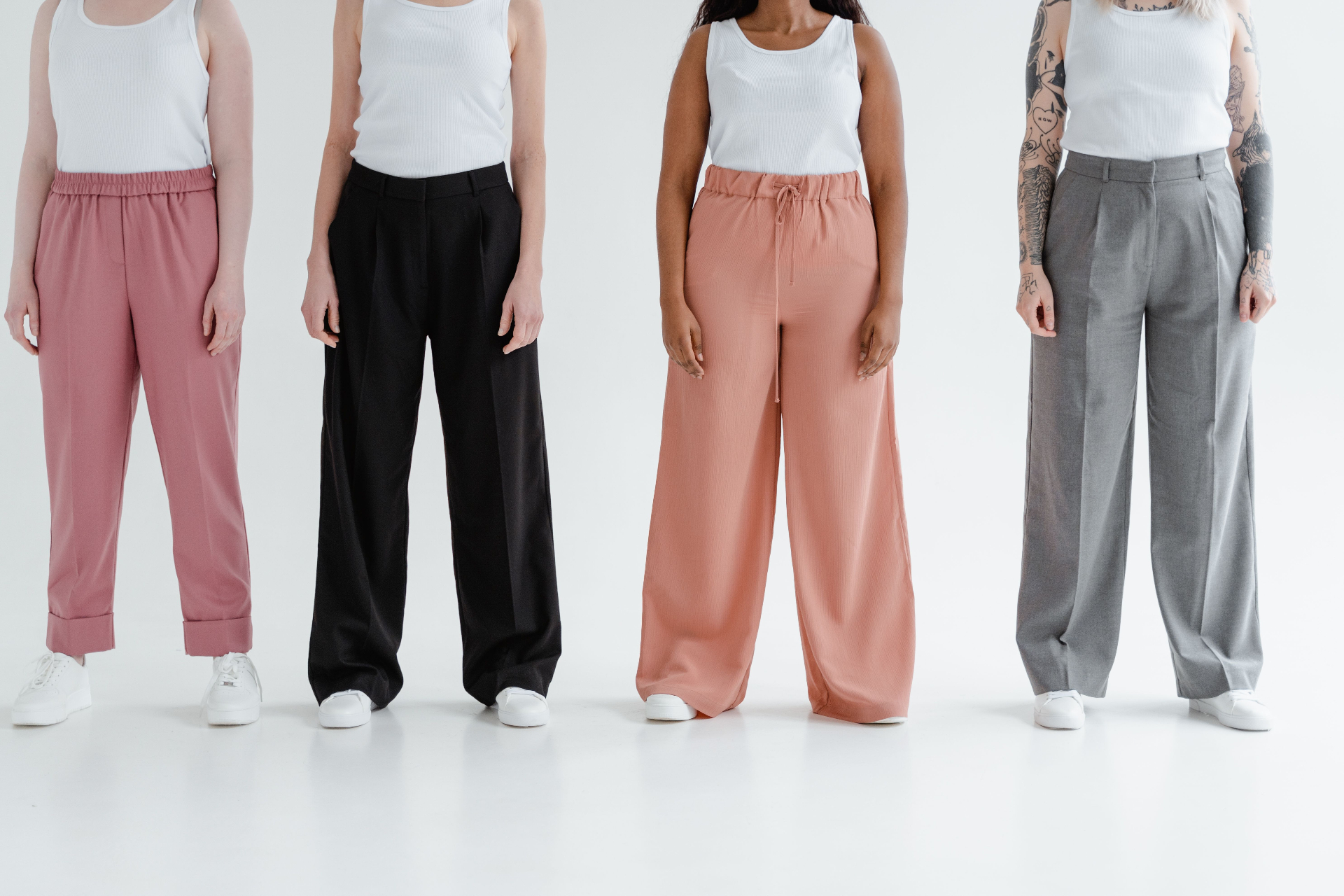 Four women in white ribbed tanks and tailored trousers in dusty pink, black, coral and grey