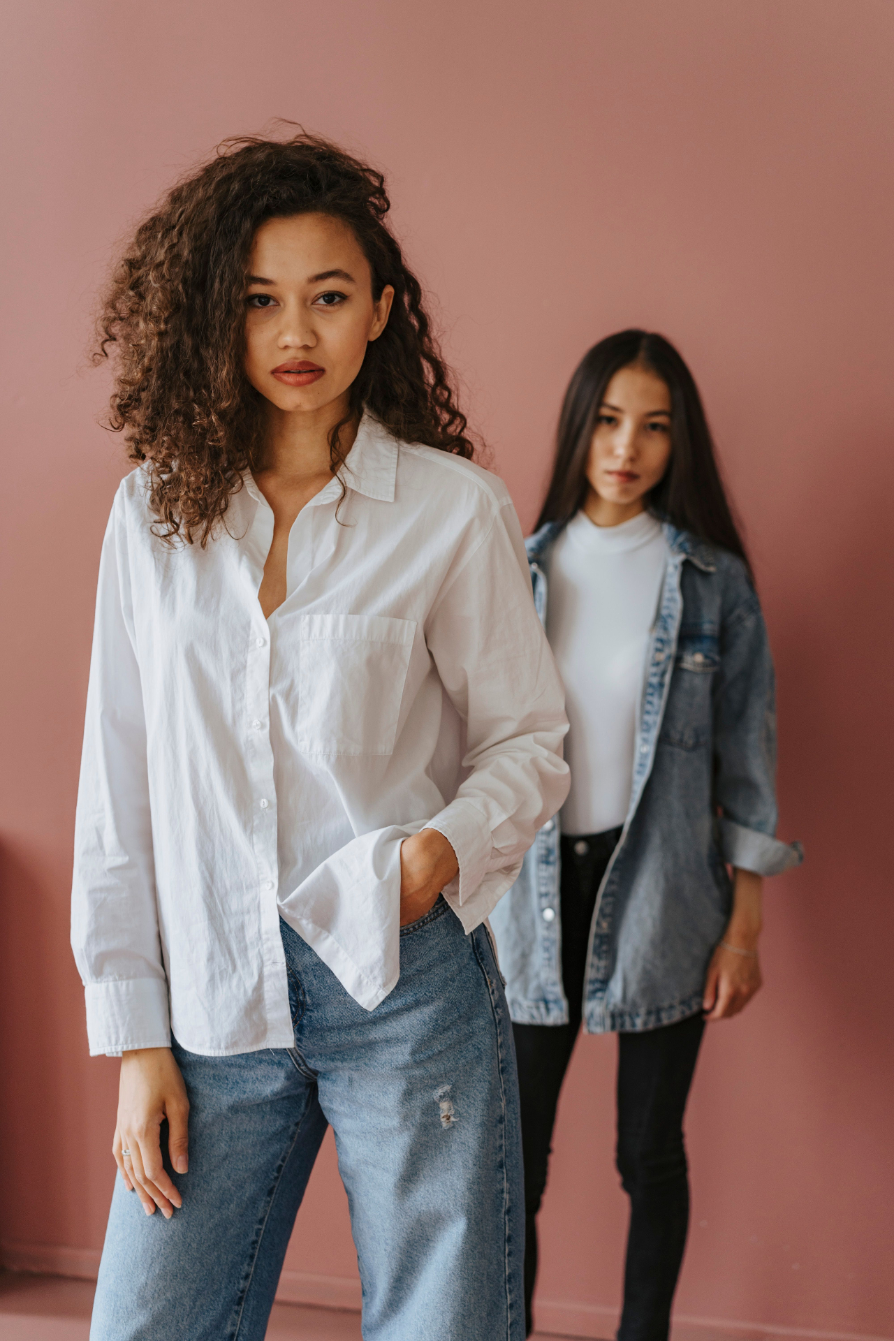 Two women in white shirts and denim, one in a relaxed white shirt with light-wash jeans, the other in an oversized denim jacket